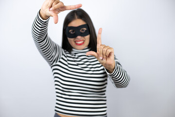 Young beautiful brunette burglar woman wearing mask smiling making frame with hands and fingers with happy face