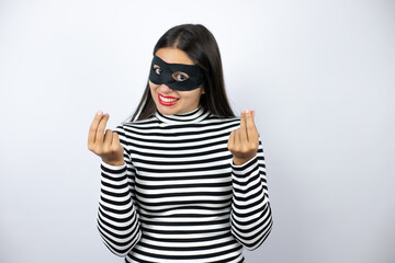 Young beautiful brunette burglar woman wearing mask doing money gesture with hands, asking for salary payment, millionaire business