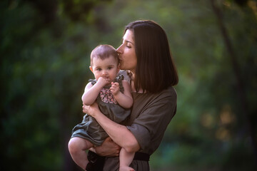 Young mother holds baby in arms in the rays of the sunset in the park. Woman in a green dress kisses, hugs a girl. Maternal care, custody. Adoption concept. Walks in the open air. Close up portrait