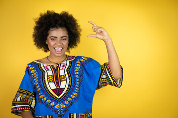 African american woman wearing african clothing over yellow background smiling and confident gesturing with hand doing small size sign with fingers . Measure concept.