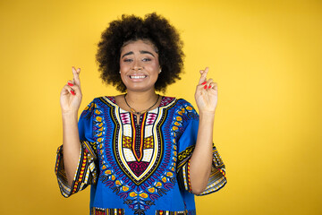 African american woman wearing african clothing over yellow background gesturing finger crossed smiling with hope and looking side