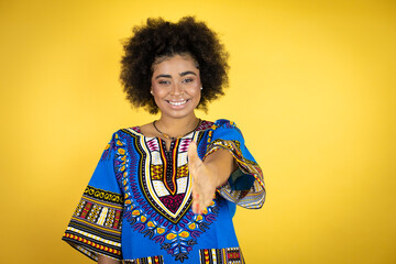 African american woman wearing african clothing over yellow background smiling friendly offering handshake as greeting and welcoming