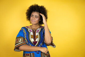 African american woman wearing african clothing over yellow background confuse and wonder about question. Uncertain with doubt, thinking with hand on head
