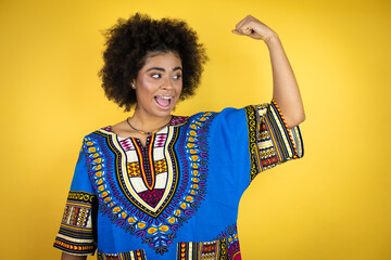 African american woman wearing african clothing over yellow background showing arms muscles smiling proud