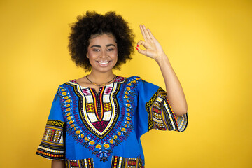 African american woman wearing african clothing over yellow background doing ok sign with fingers and smiling, excellent symbol