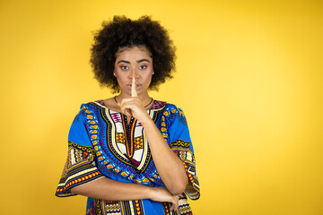 African american woman wearing african clothing over yellow background asking to be quiet with finger on lips. Silence and secret concept.
