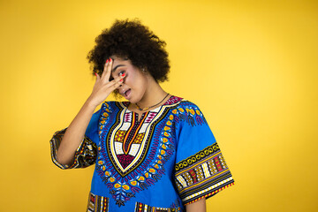 African american woman wearing african clothing over yellow background peeking in shock covering face and eyes with hand, looking through fingers with embarrassed expression