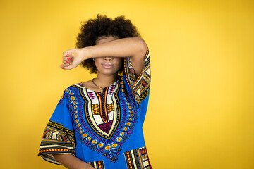 African american woman wearing african clothing over yellow background covering eyes with arm smiling cheerful and funny. Blind concept.