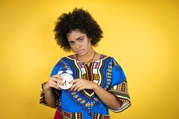 African american woman wearing african clothing over yellow background serious and holding and pointing a clock