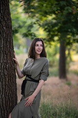 A young woman in a green khaki dress stands with her back against a tree at sunset in the park. Brunette girl smiling, walking in the garden. Close up portrait