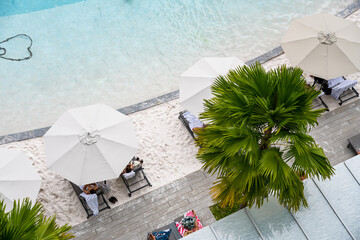Resort pool with white sand edge and cool blue water, a palm tree and umbrellas are seen from above. 