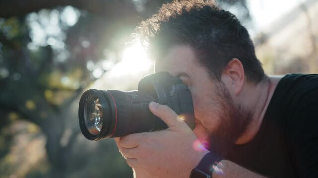 Professional photographer excited by creative photoshoot, concentrated on taking pictures in nature park on summer day at sunset. Slow motion handsome man looking in pro camera window and on screen