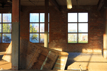 Interior view of an old, run-down, and abandoned factory building. Big, empty room with brick walls, exposed beams, and florescent light fixtures hanging form the ceiling.