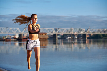Running woman at sunrise. Morning jogging on the beach or coast of river on bridge urban city background