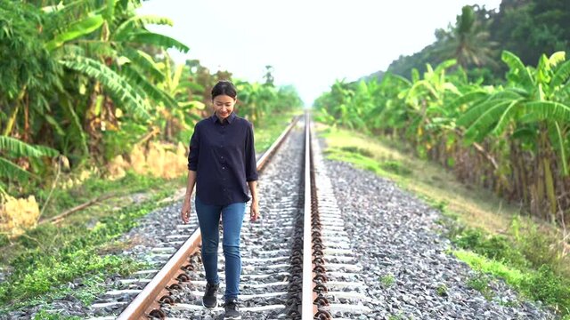 The young beautiful asian woman who tied the hair back wearing in black shirt and jean is walking and smiling happily on the local railroad looking toward the banana trees forest in the sideway