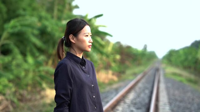 The young beautiful asian woman who tied the hair back wearing in black shirt and jean is walking and thinking about her life on the local railroad with banana trees forest in the sideway, then she st