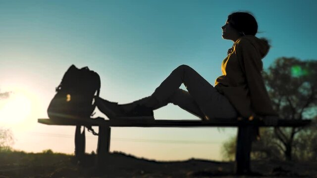 girl sitting on a bench in a park side view with a backpack resting silhouette looks at lifestyle sunset sunlight. Concept vacation relaxation teenage girl sitting back