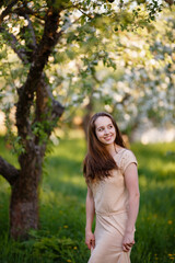 Young woman in beige dress in a blooming apple garden. Spring story. Brown-haired girl with long hairs. Woman plays with her hairs