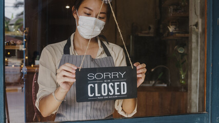 Young Asia girl wear face mask turning a sign from open to closed sign on glass door cafe after...