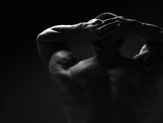 Strong muscular man, athlete, sportsman is standing naked, shirtless with his back to camera holding hands behind head over dark background