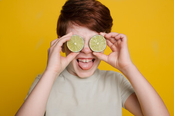 Caucasian boy is covering his eye with sliced lime posing on a yellow studio wall