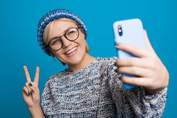 Charming woman is making a selfie of herself gesturing peace sign at camera while wearing glasses...