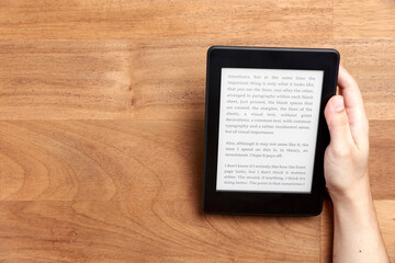 Young hand holding an electronic reader with text on the screen on a wooden desk