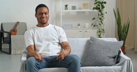 Afro american young sad guy black ethnic man in white t-shirt sits on gray sofa in cozy living room watching football or news program on TV aggressively waving his hands throwing remote controller