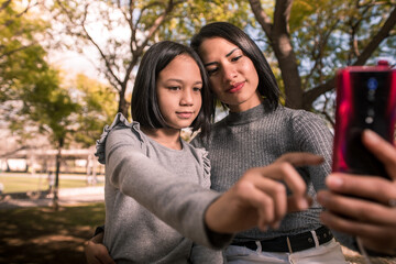 Young mother and her daughter taking a photo with the phone at the park on a sunny day. Little girl touching the smartphone.