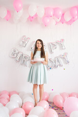 Brunette woman in mint dress holding pink cake with candles. Birthday decorations with white and pink color balloons and confetti for party on a white wall. Happy birthday 30th anniversary, 30 years.