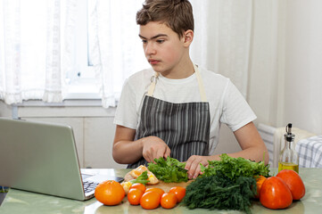 A teenage boy prepares a fruit salad on a recipe from the Internet. The boy cuts lettuce on the kitchen table, in front of him is a laptop. The child is engaged in household chores.