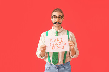 Young man with in funny disguise holding poster with text APRIL FOOLS DAY on color background