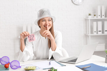 Young businesswoman holding sticky note with text KICK ME at workplace in office. April Fools Day celebration