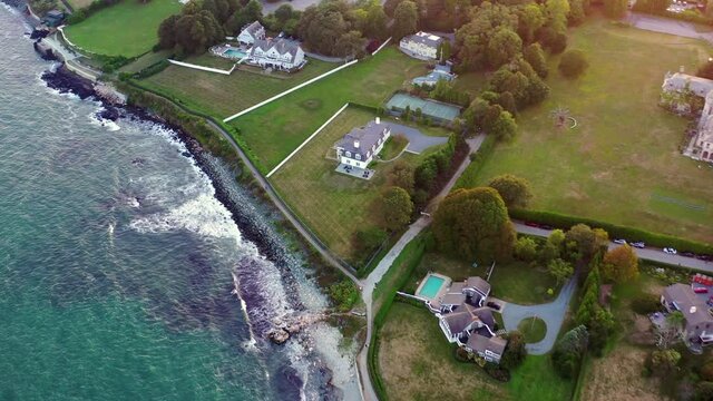 Aerial panning shot of houses by waves splashing on coastline, drone flying forward over city at sunset - Newport, Rhode Island