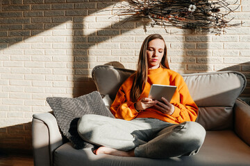 Pretty young woman with tablet sitting on sofa at home. Bright yellow orange sweatshirt and gray...