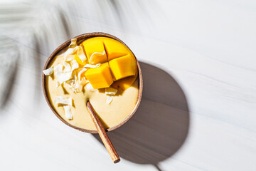 Mango smoothie bowl with coconut flakes in coconut shell, white marble background. Vegan food concept. Healthy sugar free dessert.