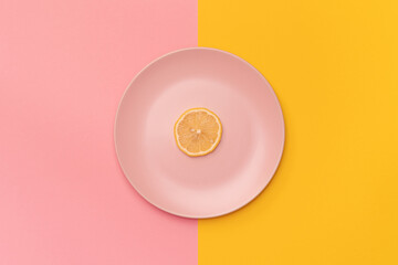 Slice of the fresh lemon on a pink plate. The background is divided vertically into pink and yellow. The lemon in the center of the frame. Minimalism