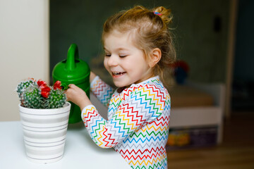 Little toddler girl watering flowers and cactus plants on window at home. Cute child helping,...