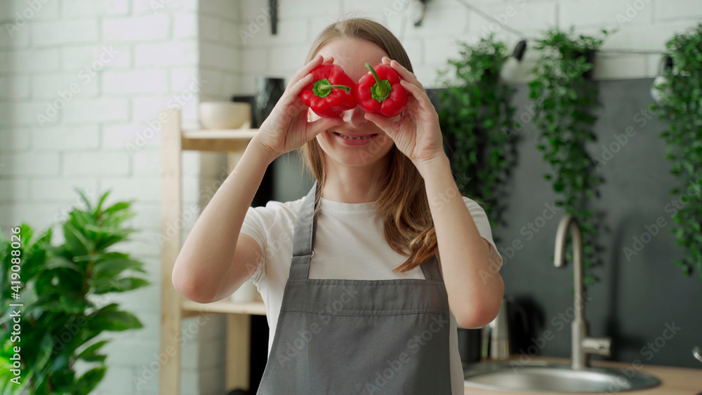 Wall mural happy young woman smiles and has fun playing with red pepper in front of her eyes in the home kitche