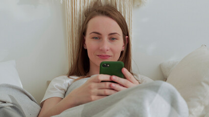 Smiling woman using a mobile phone was lying on the bed. Happy brunette young woman uses mobile phone at home.