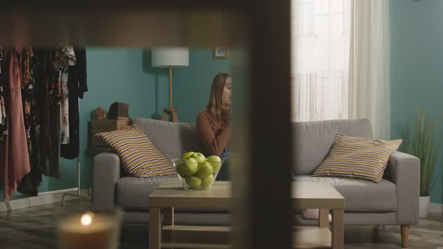 Young lady in blue jeans and brown sweater sits on beige sofa on window background, bites red apple and talks on video call. On coffee table is glass jar with green apples. Camera dollies horizontally