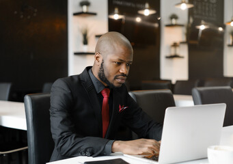 An image of an African-American businessman, dressed in a black suit, working on his laptop. A handsome young man at a desk in an office with a black and white interior