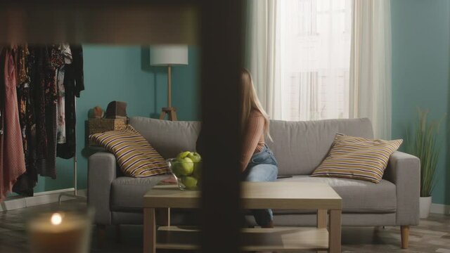 Young girl in blue jeans and brown sweater sits on beige sofa on window background, bit red apple and choked. Trying to cough. On table is glass jar with green apples. Camera dollies horizontally