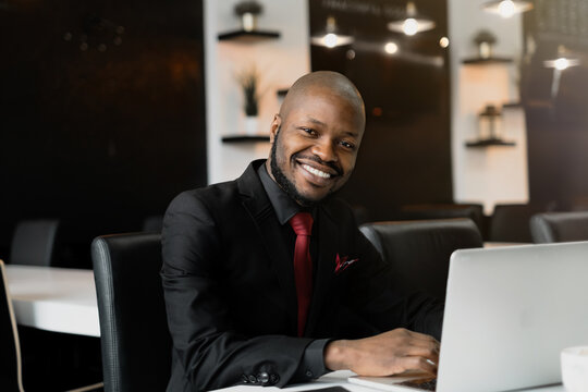 An image of a smiling African-American businessman, dressed in a black suit, working on his laptop. A handsome young man at a desk in an office with a black and white interior