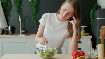 Unhappy woman eating vegetable salad at table in kitchen. Displeased young woman eating green leaf...