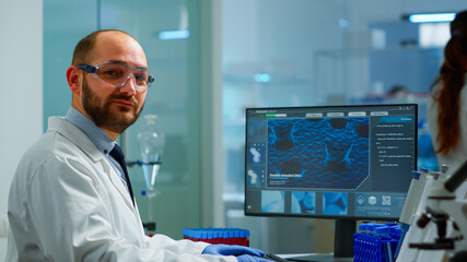 Portrait of scientist man looking at camera sitting in modern equipped laboratory. Scientist doctor examining virus evolution using high tech typing on computer chemistry tools for scientific research