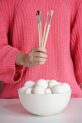 Girl in pink sweater holding paintbrushes ready to paint many eggs in bowl for Easter. Easter concept