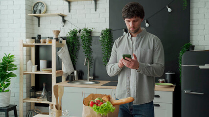 Man stands in the kitchen near a paper bag full of fresh food and uses a smartphone app to deliver it to the supermarket