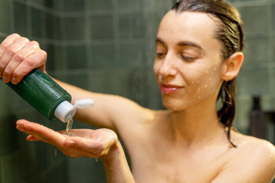 Woman Takes Shampoo From Green Bottle, Taking Shower In Green Bathroom With Water Dripping On Face. Close Up. Wet Hair Combed Back.