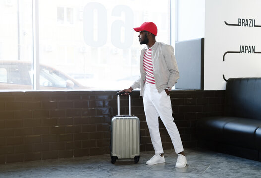 Portrait of an African-American man wearing a white jacket, white pants, a red and white T-shirt and a red baseball cap, with a suitcase going on vacation, waiting for his flight